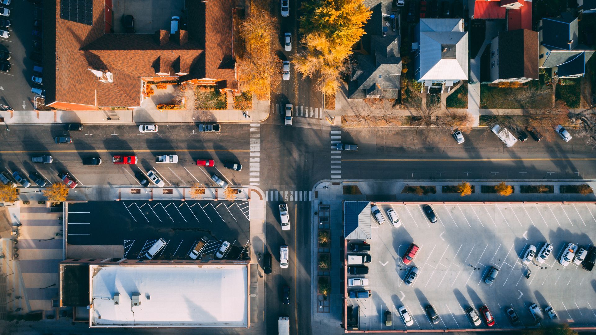 An aerial view of a town intersection with parked cars, buildings, and tree-lined streets during the day.