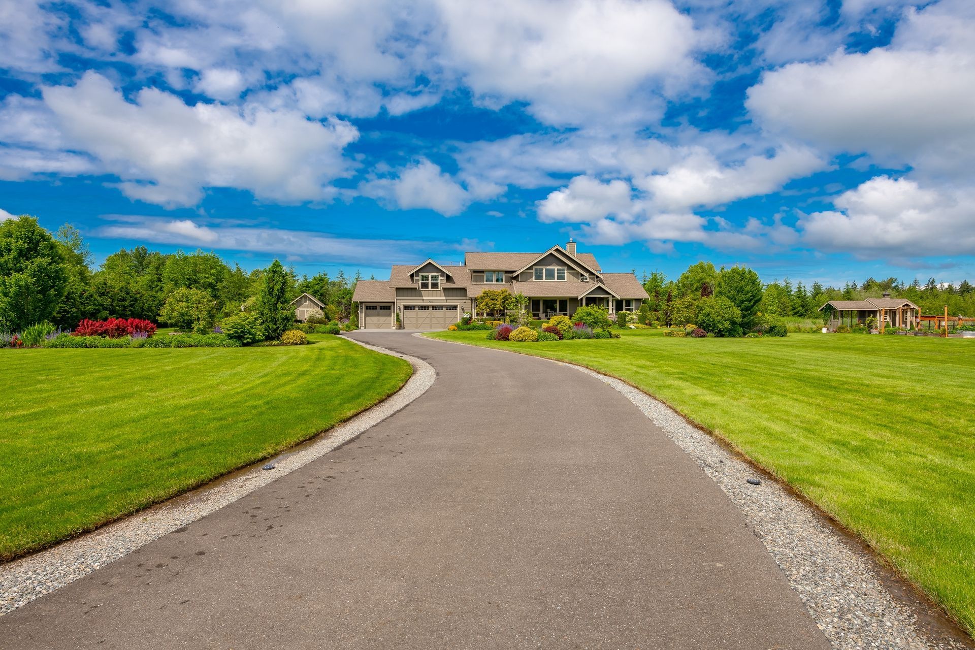 A curved asphalt driveway leads to a large, multi-story house surrounded by a lush green lawn and trees under a blue sky.