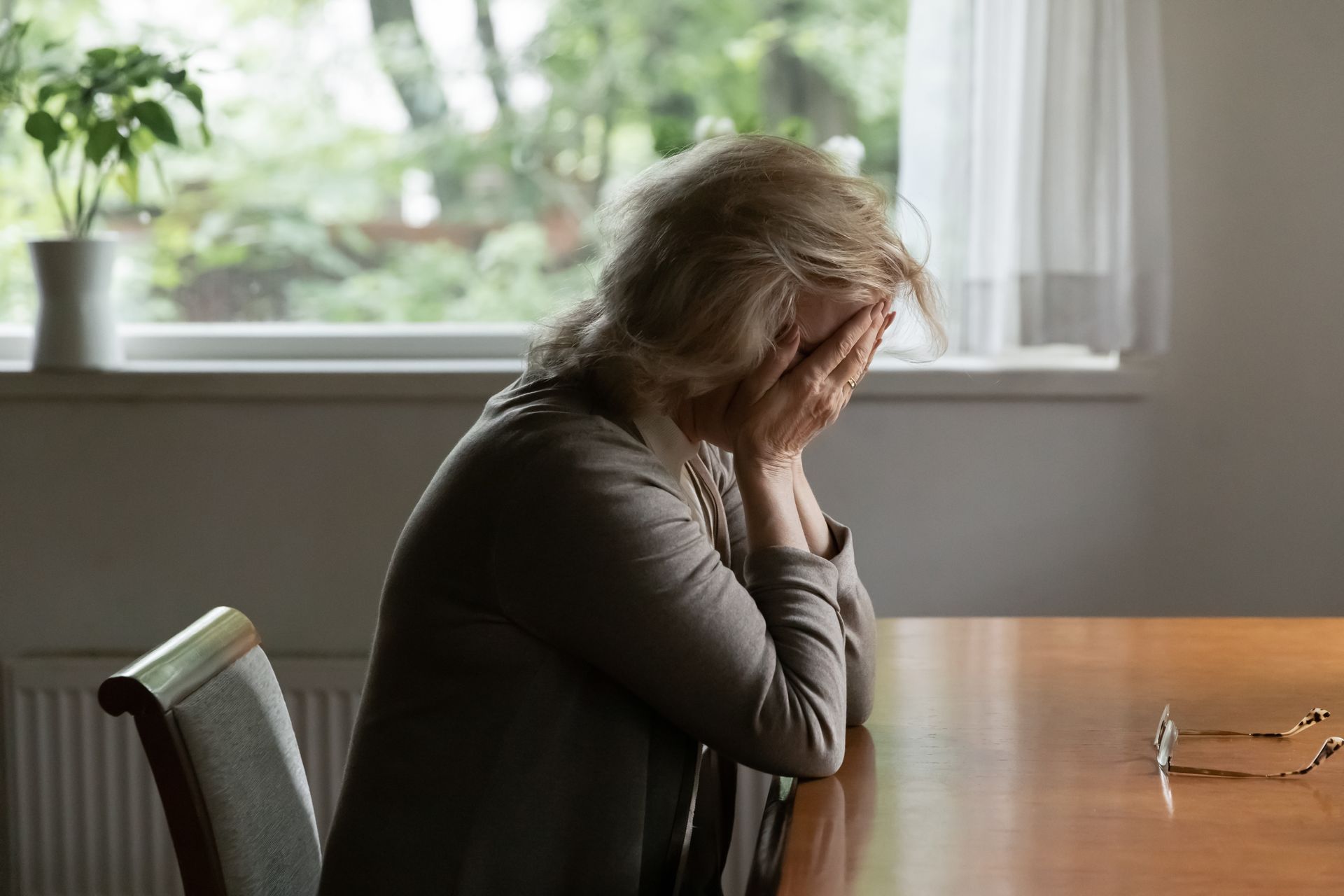 A person sits at a wooden table with their head in their hands, appearing distressed or exhausted in a brightly lit room.