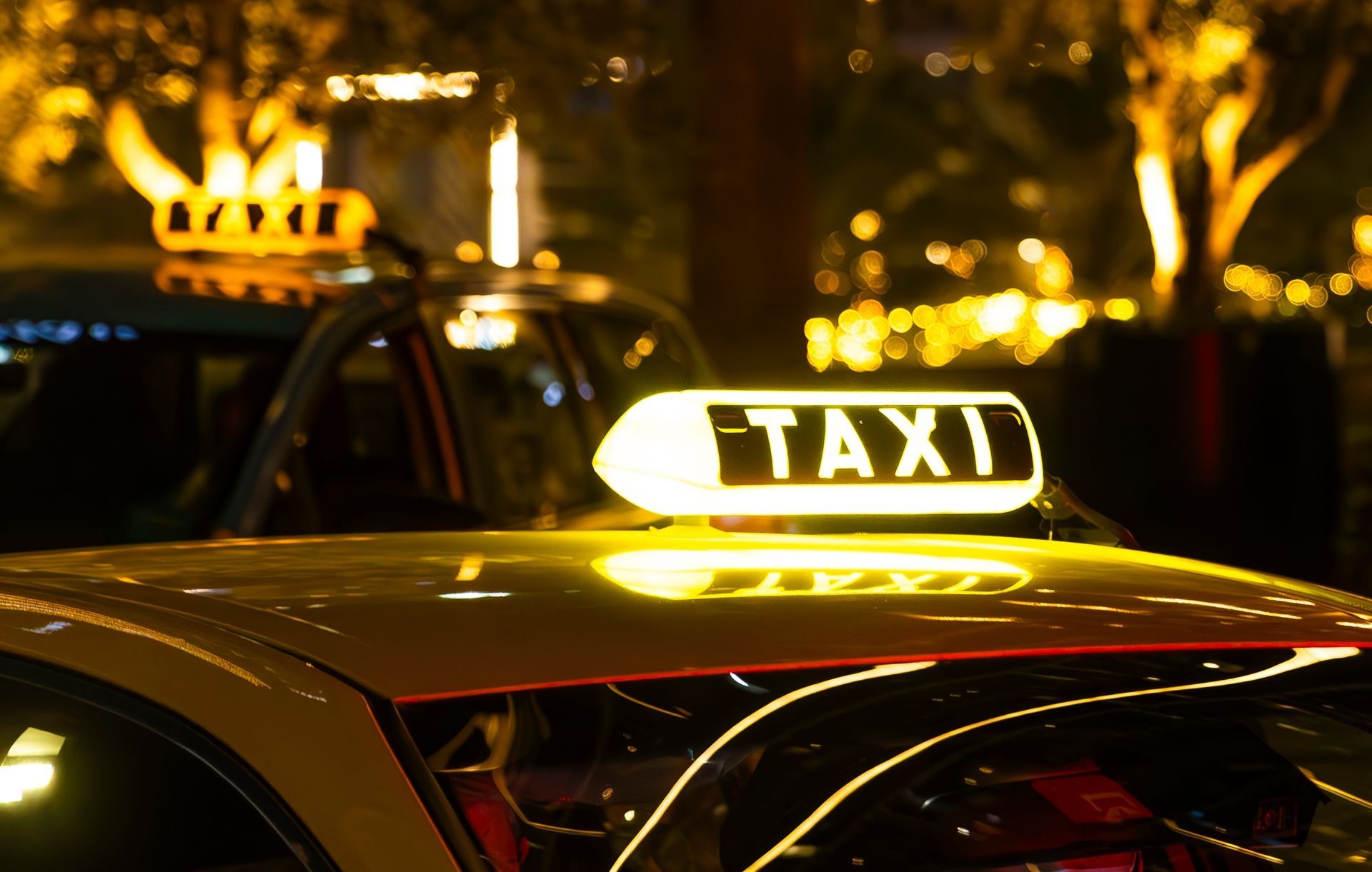 Close-up of a glowing yellow taxi light on a car roof at night, with another taxi blurred in the background.