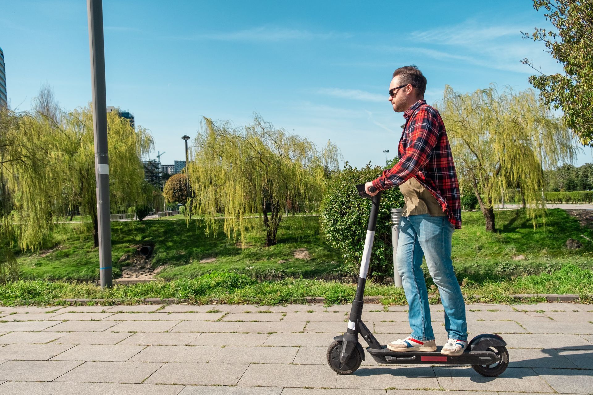 A person wearing a plaid shirt and jeans riding an electric scooter along a sunny outdoor path.