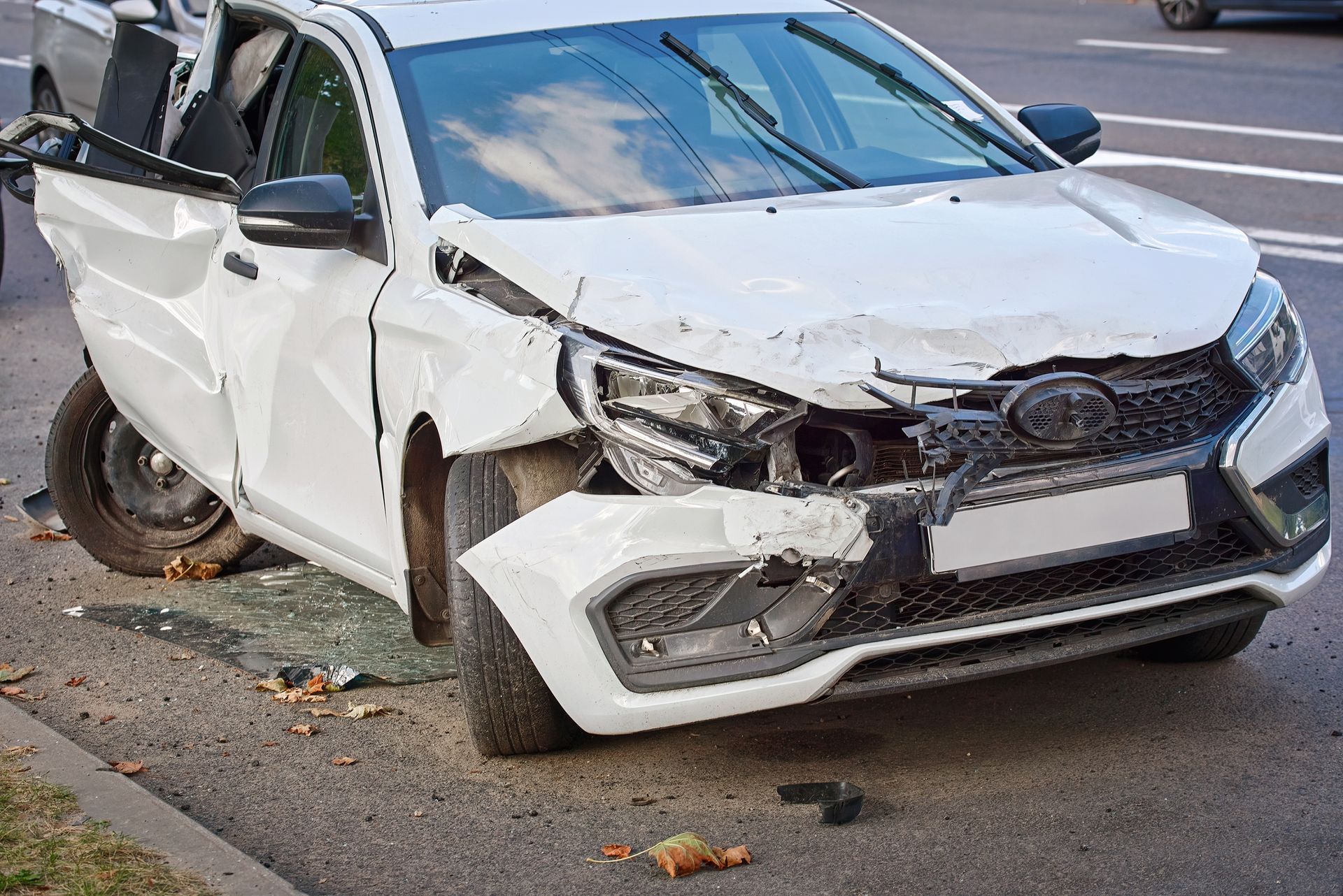 A heavily damaged white sedan sits on the side of a road, showing significant front-end and side collision damage.