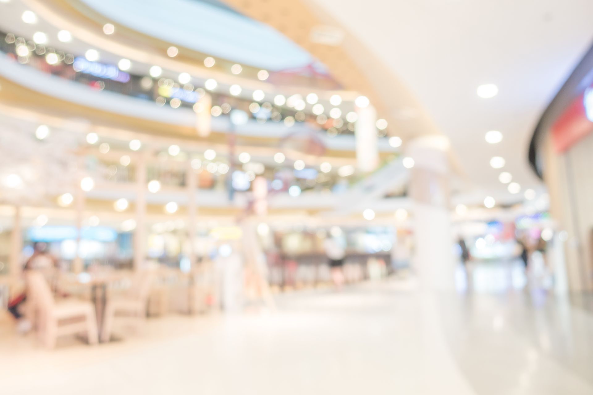 A blurred, bright, multi-level indoor shopping mall with warm lighting and shoppers in the distance.