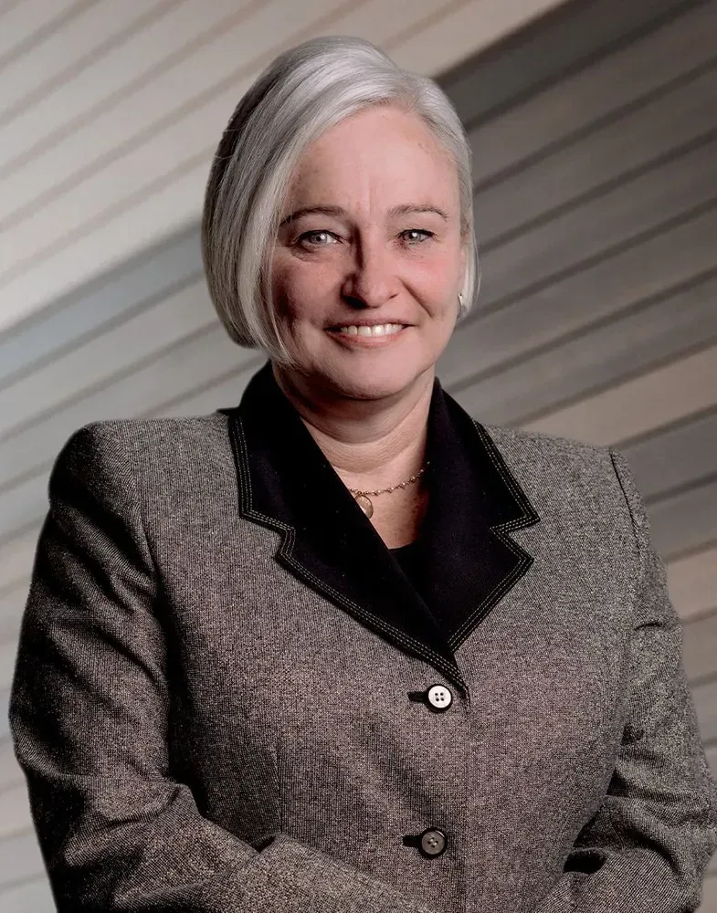 Smiling woman with short gray hair in a tweed blazer, black collar, and gold necklace, set against a light-colored, modern backdrop.