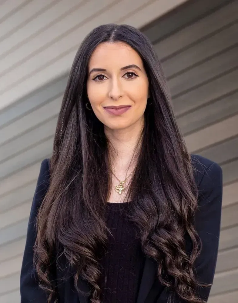 Woman with long dark hair, wearing a black blazer and top, smiling with a neutral background.