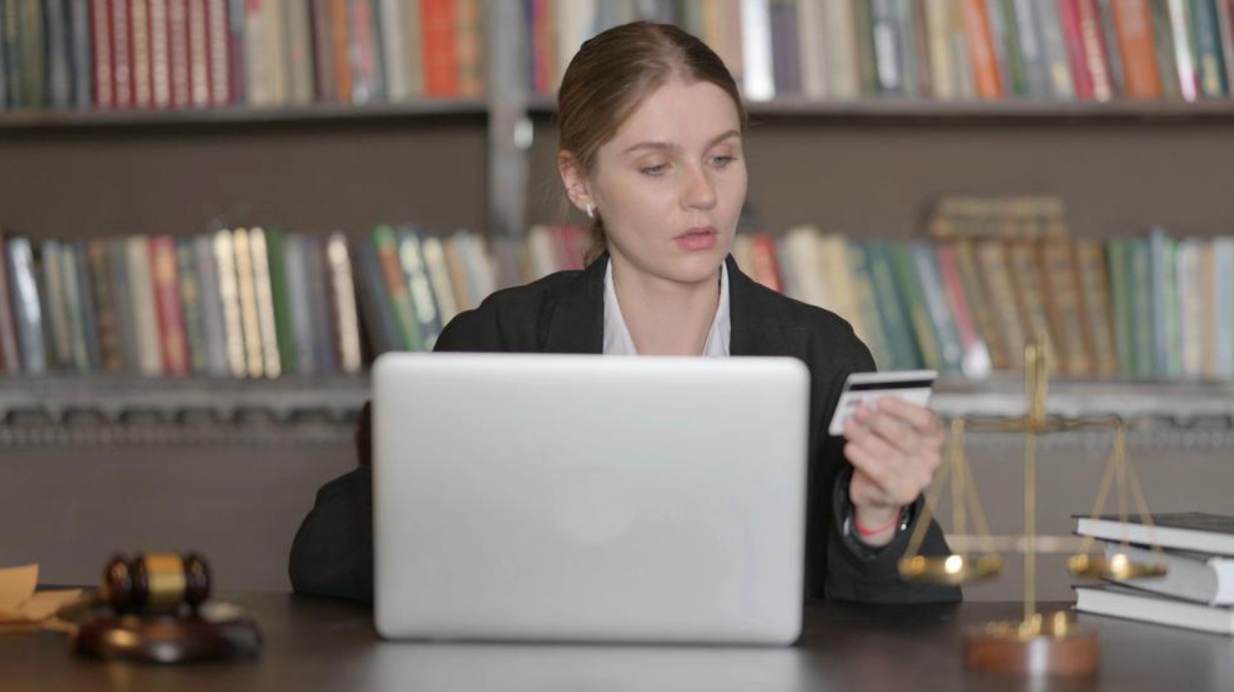 A woman is sitting at a desk in front of a laptop computer holding a credit card.