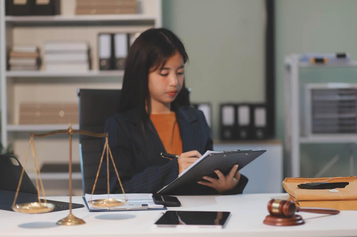 A woman is sitting at a desk in an office looking at a clipboard.