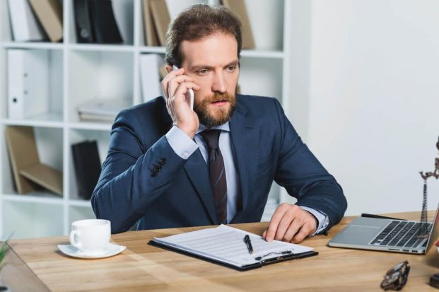 A man in a suit and tie is sitting at a desk talking on a cell phone.