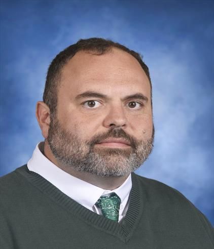 A person with a beard wearing a green sweater and tie, centered against a blurred blue background.