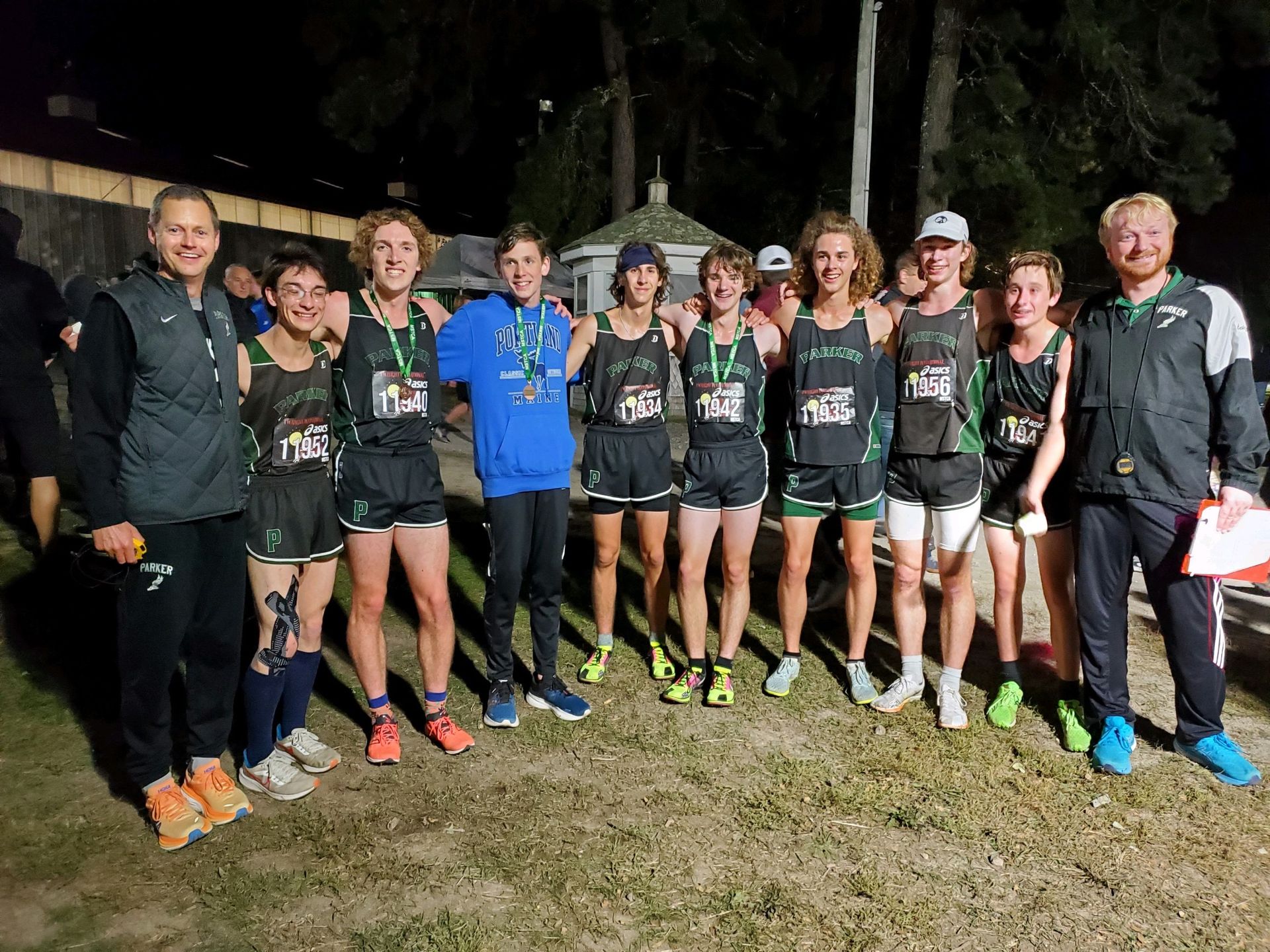 A group of cross-country runners in matching uniforms standing outdoors with their coaches at night.