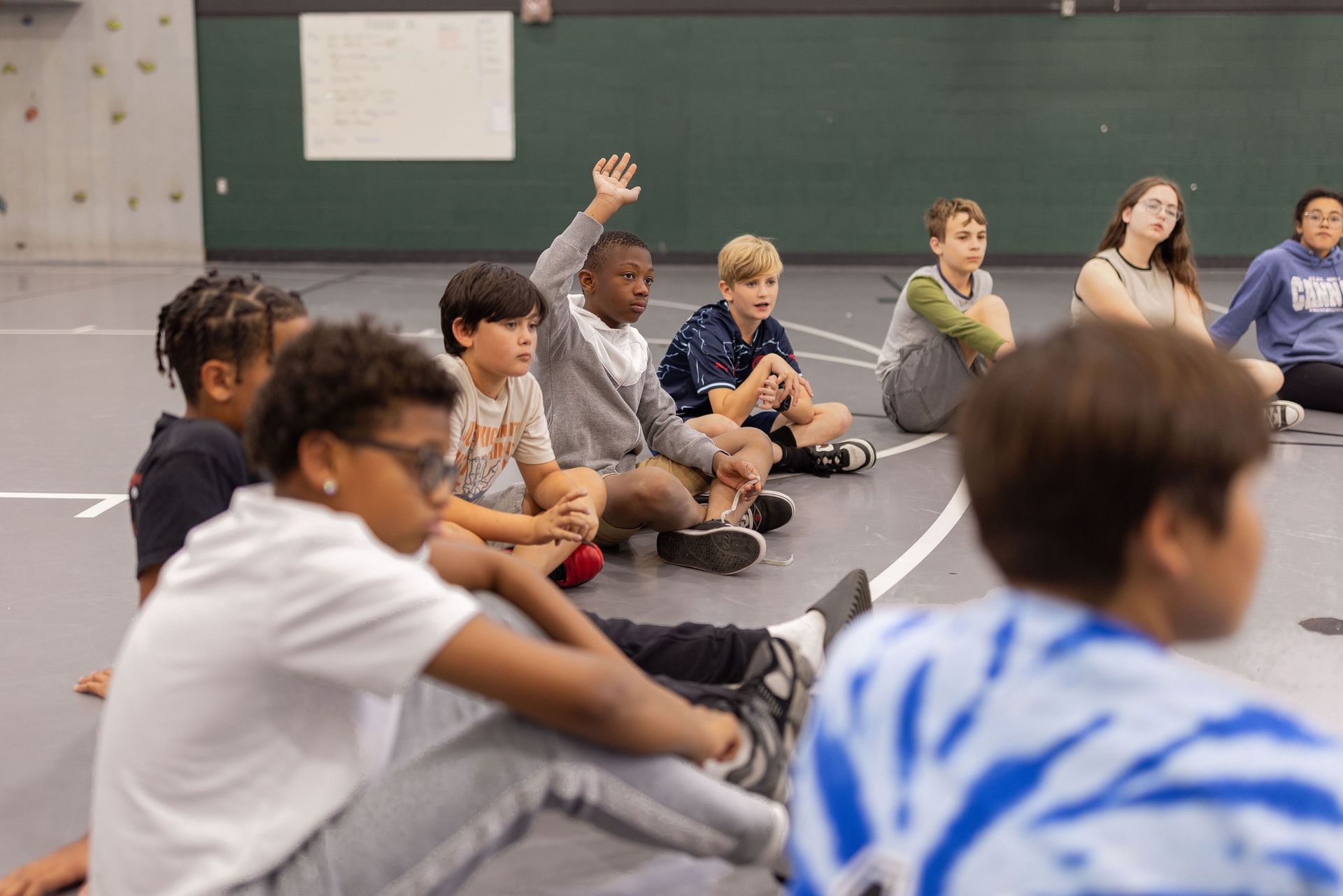 A group of students sits on a gymnasium floor in a circle, with one student raising their hand.