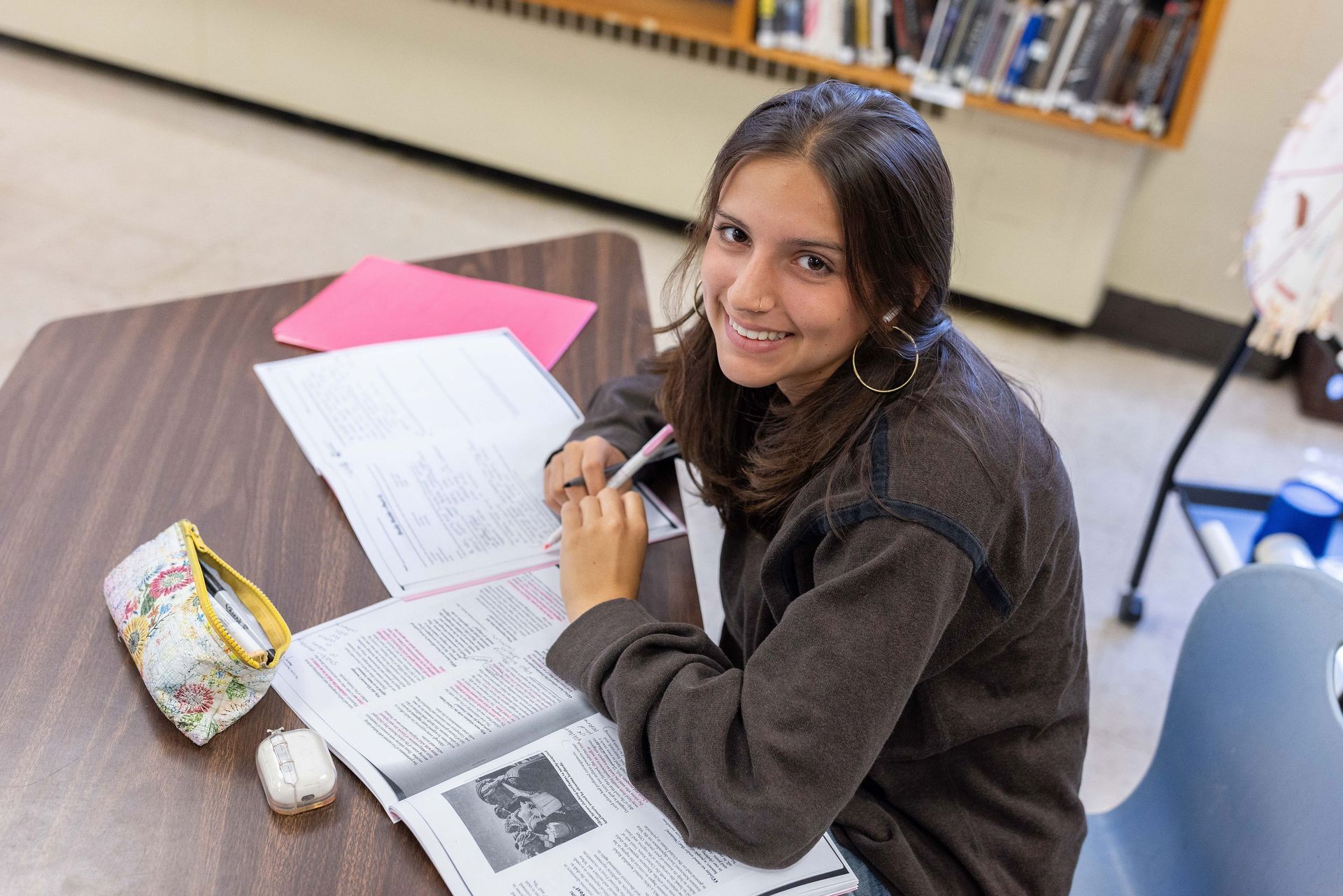 A person sitting at a library table, smiling while writing in a notebook next to a pencil case and a pair of earbuds.