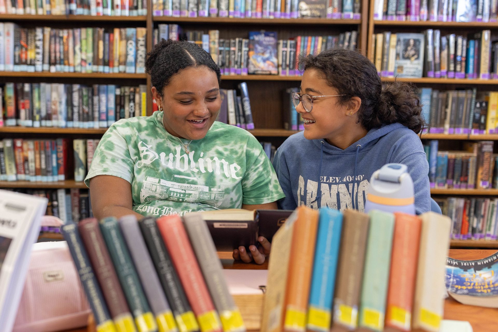 Two people smile while reading a book together in a library, surrounded by shelves full of books.