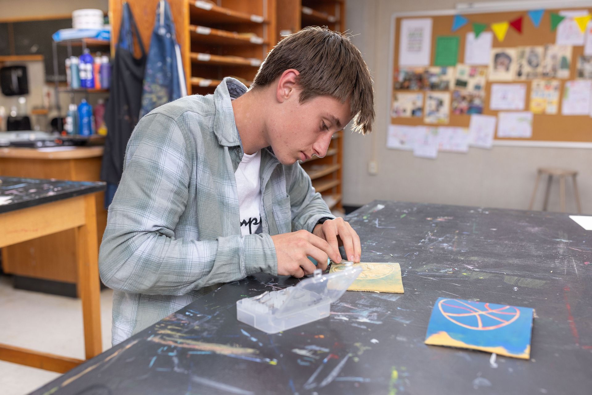 A person in a plaid shirt sits at a dark table, focused on carving a small, rectangular block in an art classroom.