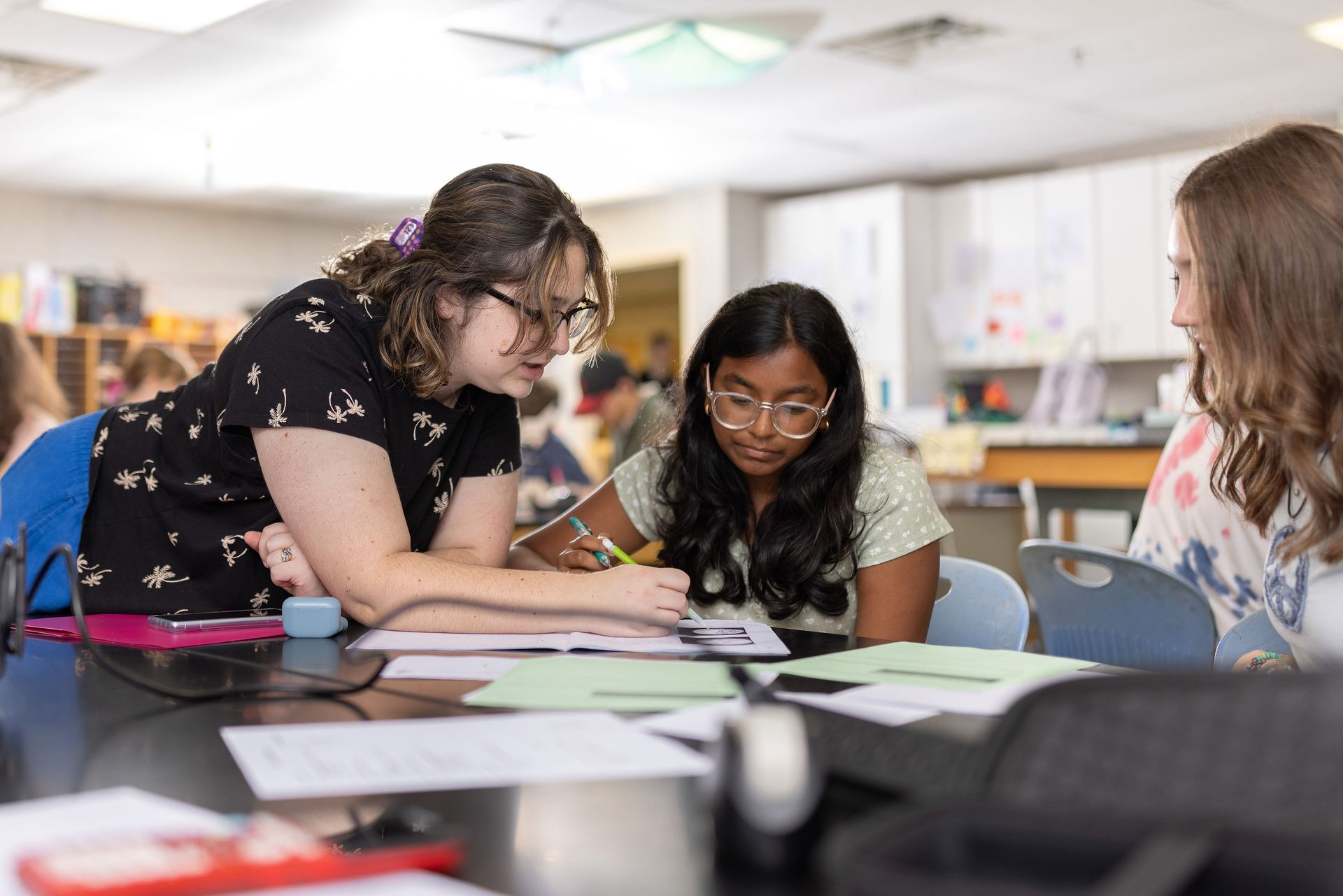 Three students collaborate at a desk in a classroom, focused on a document while one person leans in to provide guidance.