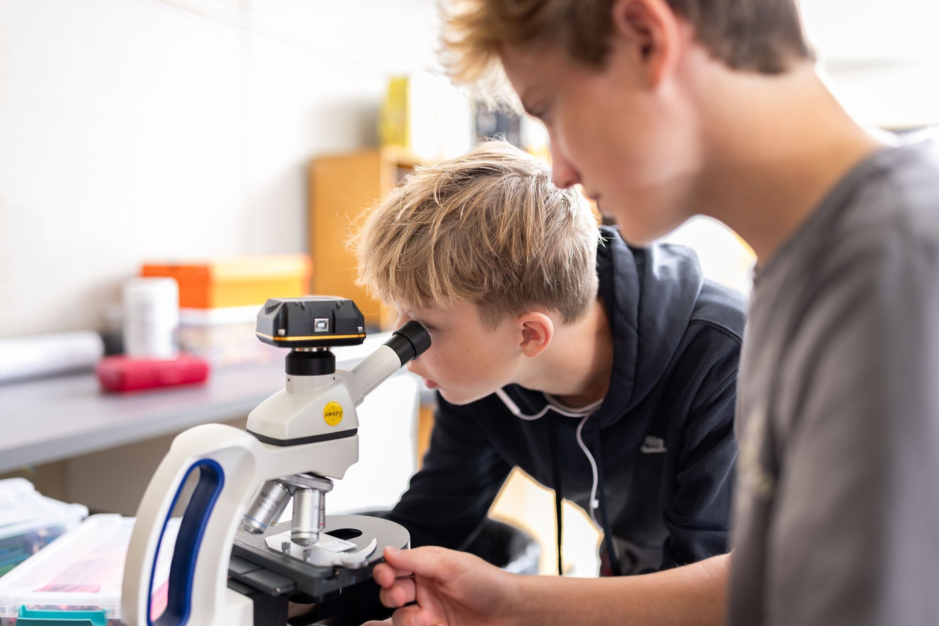Two students use a microscope to study samples in a classroom setting.