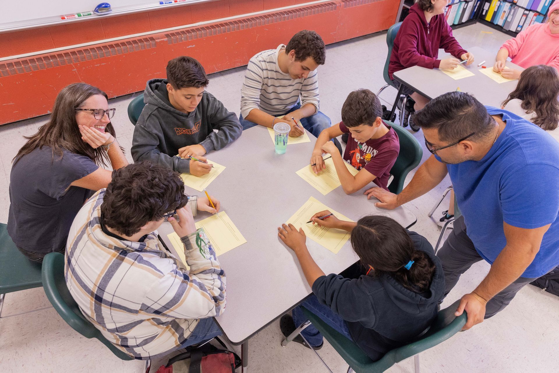 A teacher stands at a square table in a classroom while a group of students works on papers, some engaged in discussion.