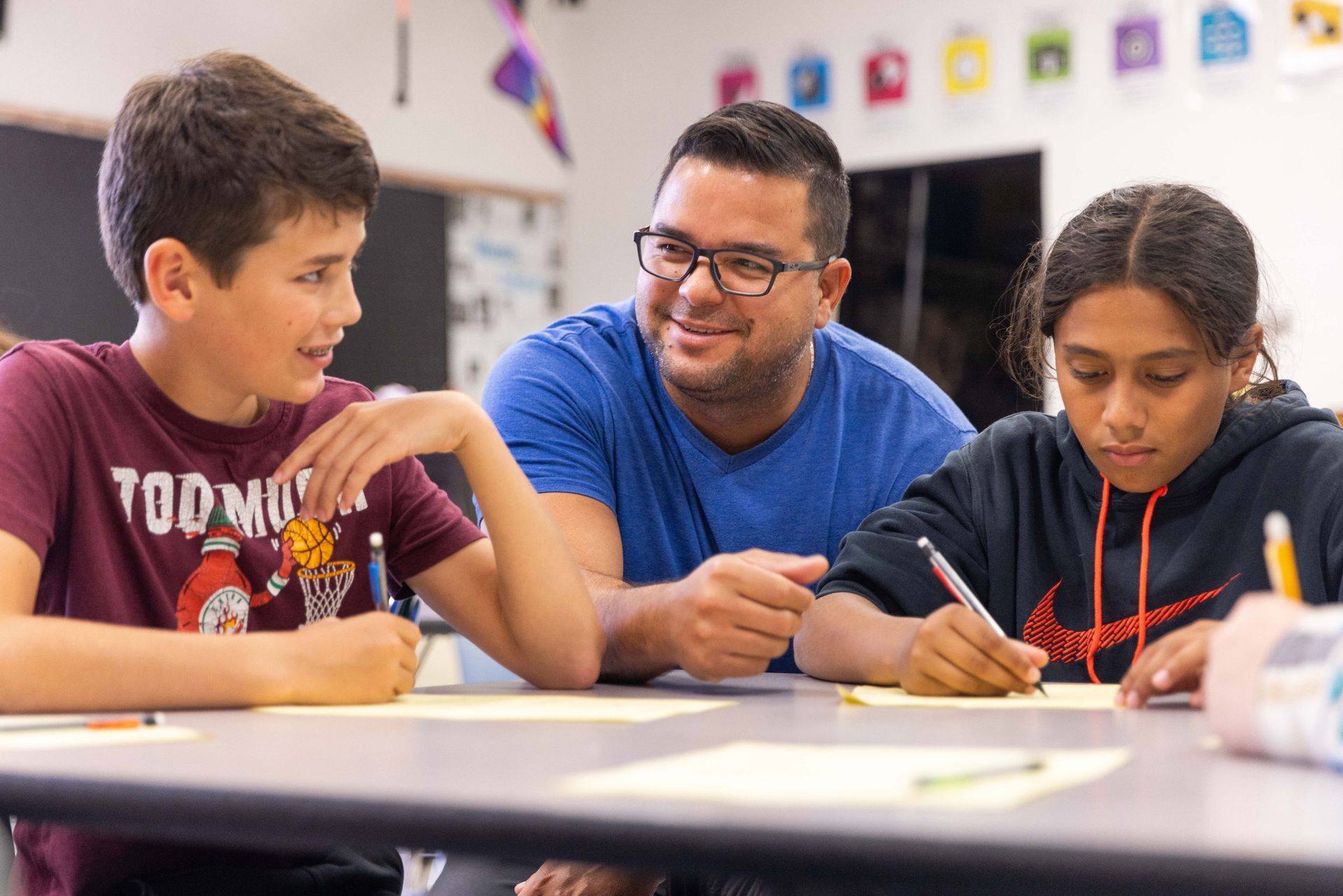 A teacher sits at a desk with two students, assisting them as they work on a classroom assignment.