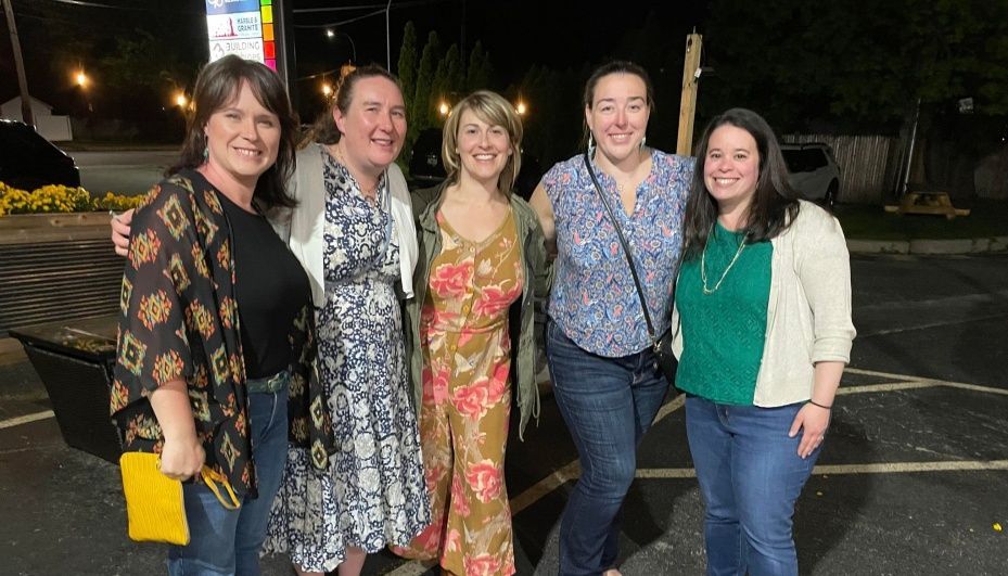 Five people stand smiling together in a parking lot at night, dressed in casual clothing.