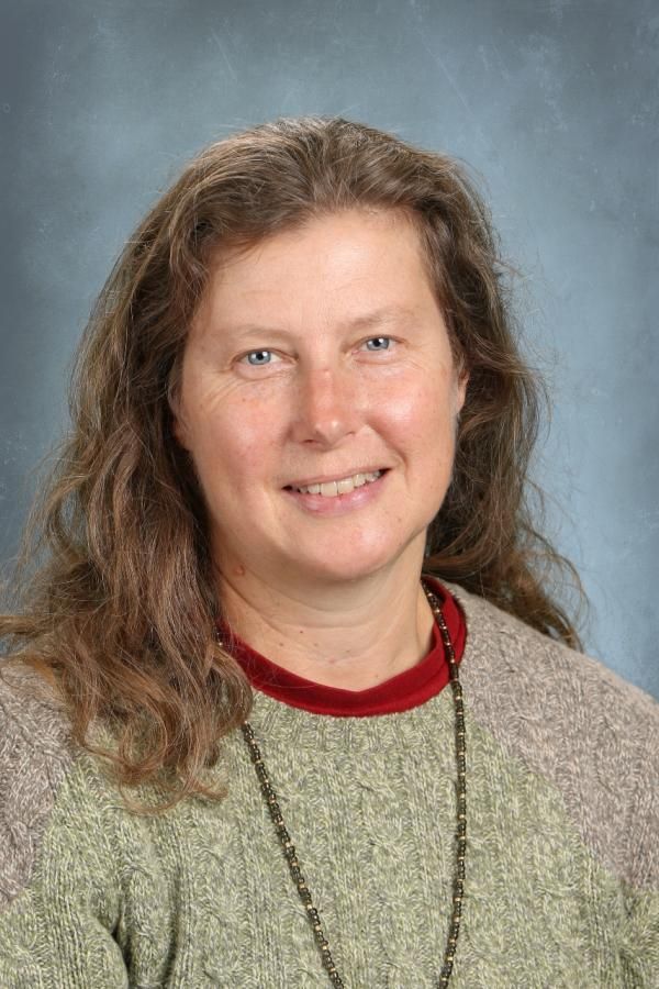 A person with long brown hair smiling against a blurred blue background, wearing a textured olive sweater and a necklace.