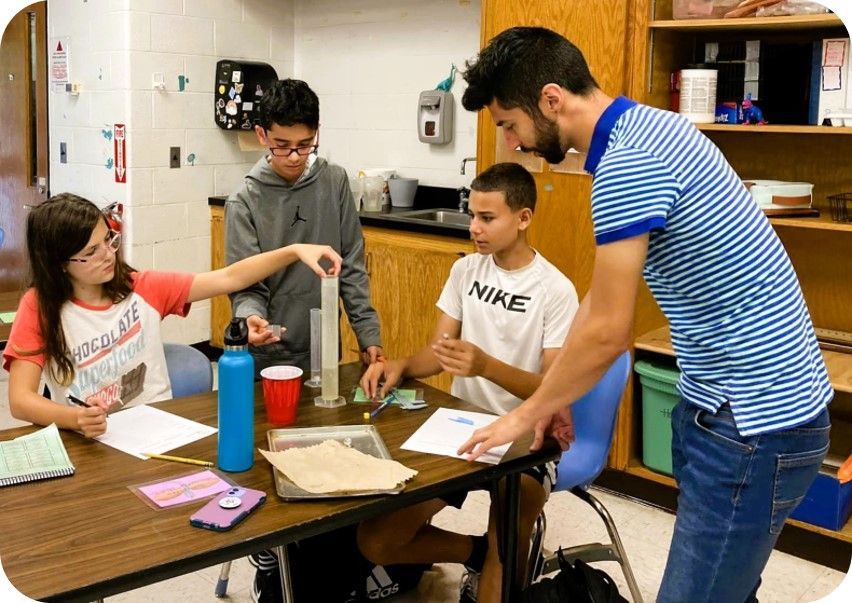 A teacher assists three students at a table in a classroom as they conduct a science experiment with a graduated cylinder.