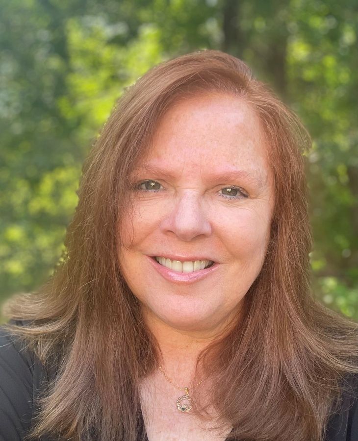 A headshot of a smiling person with shoulder-length reddish-brown hair, wearing a necklace against a blurred green background.