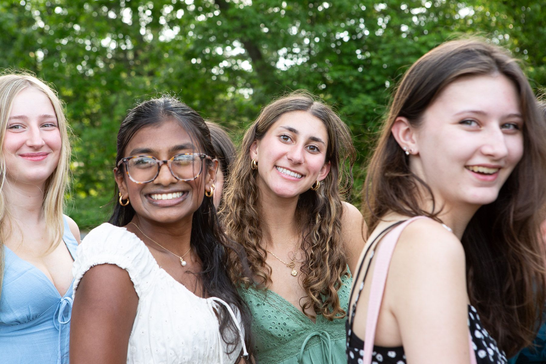 Four smiling individuals stand together outdoors in front of green trees, wearing casual summer tops.