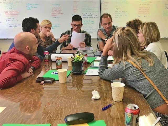 A group of people sit around a table in a meeting, with some looking at documents and others engaged in discussion.