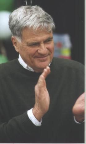 A man with graying hair wears a dark sweater and white shirt, smiling and clapping his hands together.