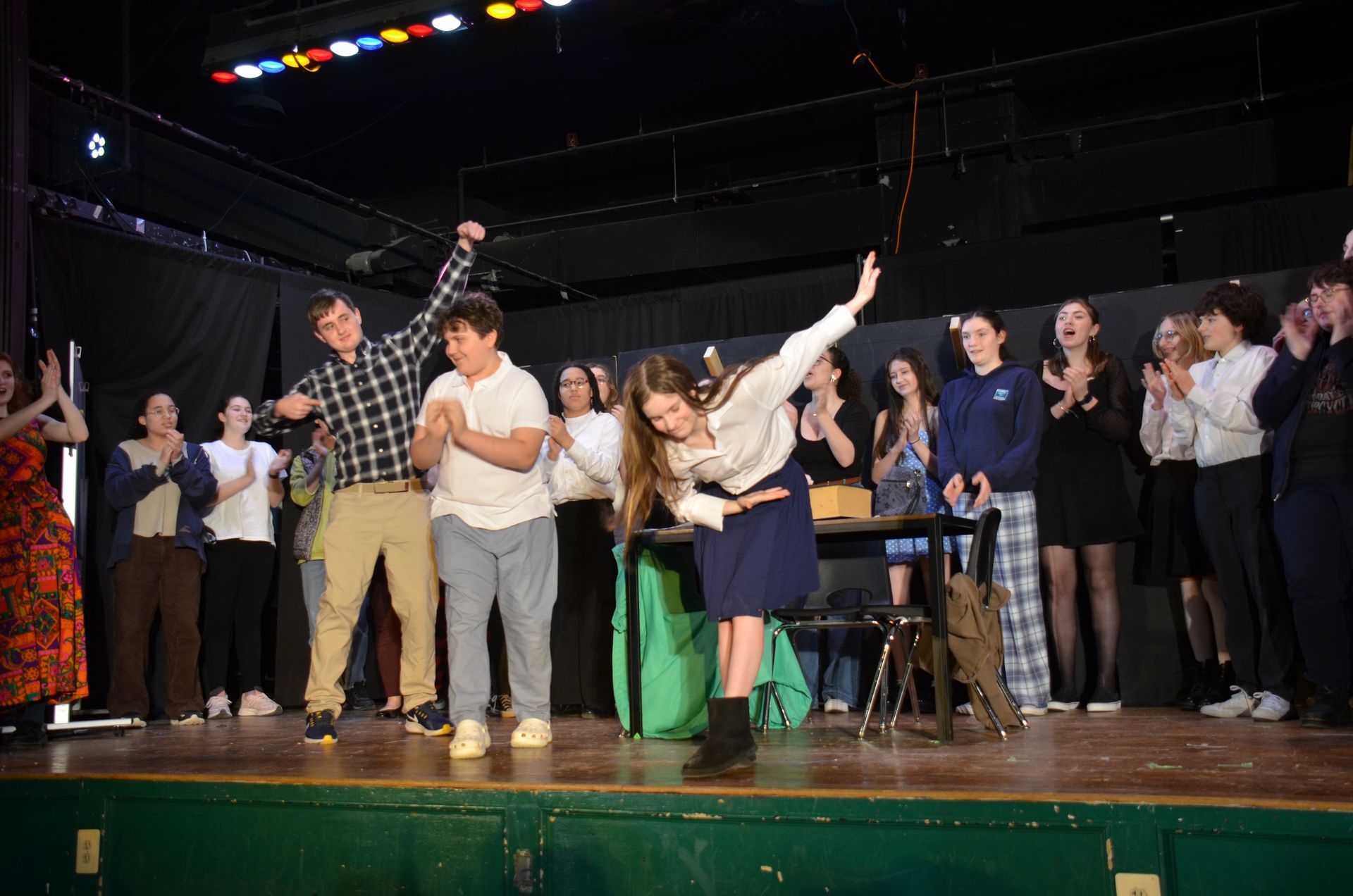 A group of people on a theater stage, with several individuals bowing or gesturing toward the audience after a performance.