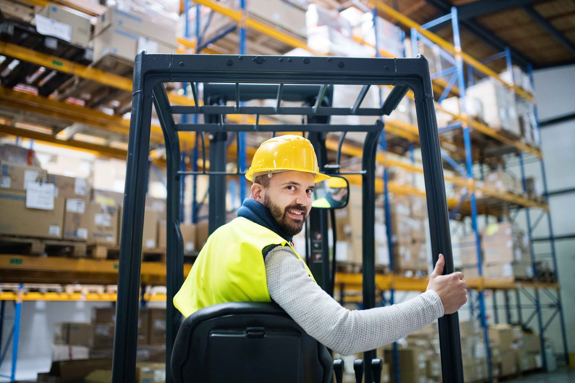 Warehouse worker in yellow hard hat and vest, giving thumbs up while operating a forklift.