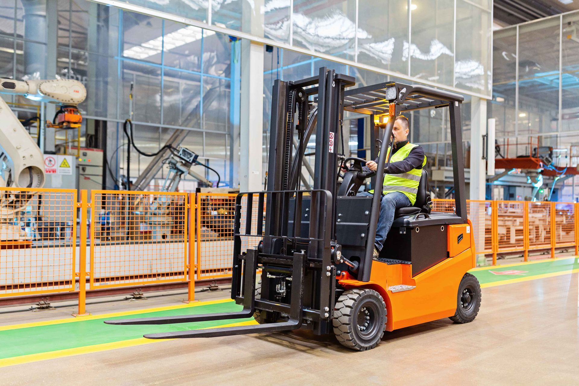 Forklift operator wearing a vest in an industrial facility. The orange forklift moves past a yellow line.