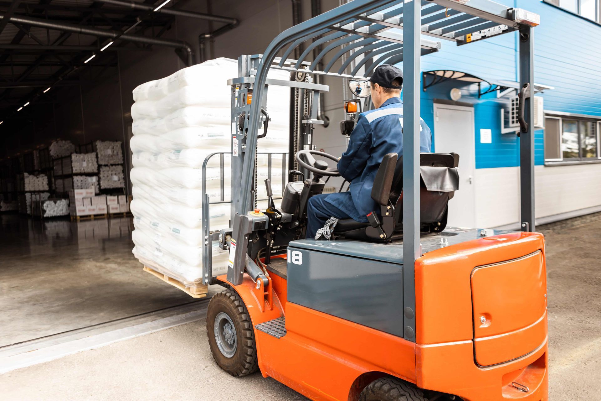 Forklift with driver carrying a pallet of white bags, near a warehouse door.
