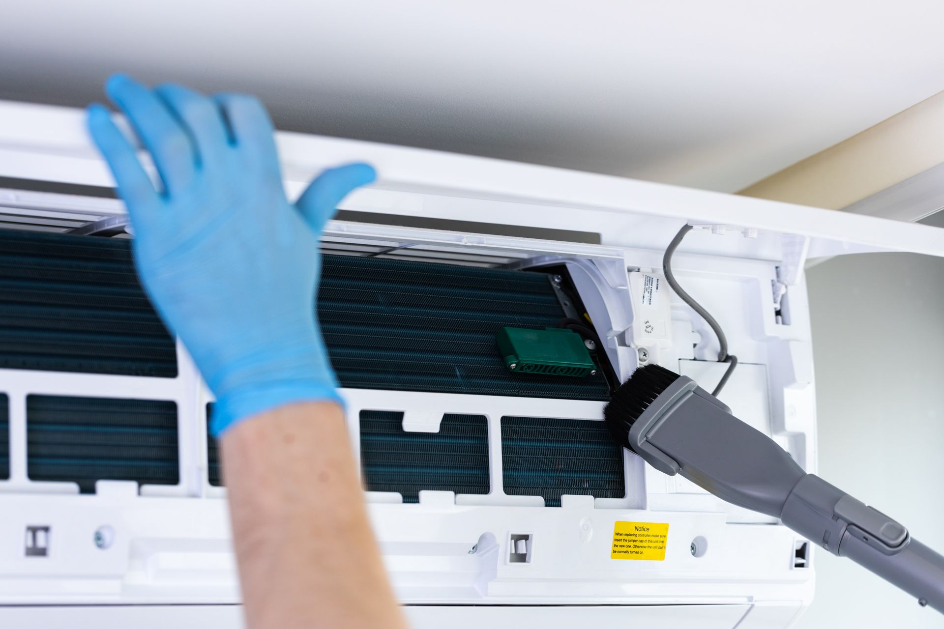 A person wearing blue gloves vacuums the interior cooling fins of a white wall-mounted air conditioner.