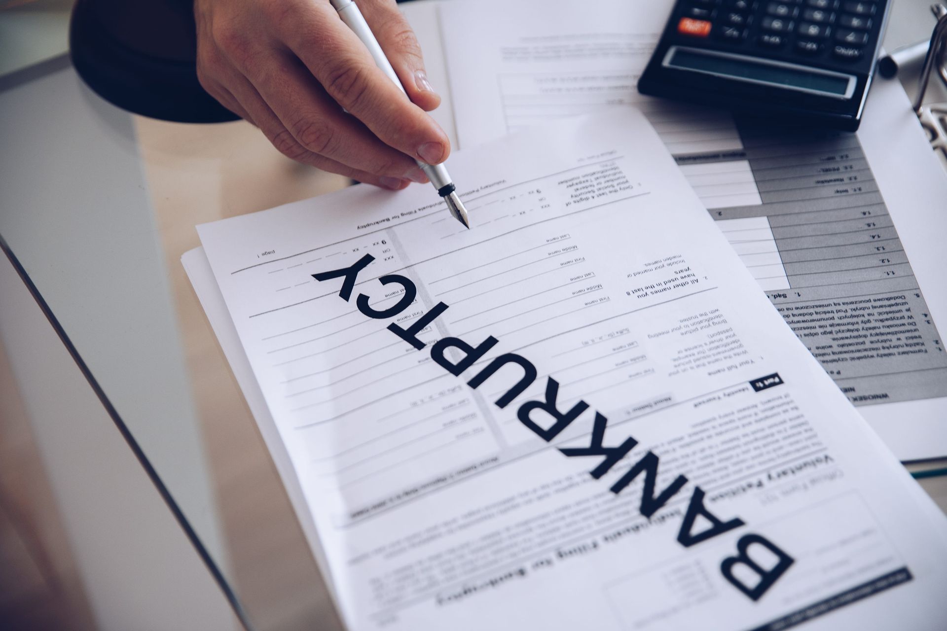 Hand holding a pen over bankruptcy paperwork on a desk with a calculator and receipt tray