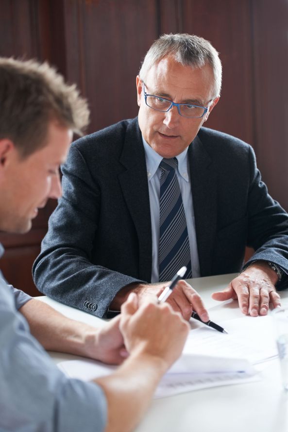Two men in a meeting, one speaking and pointing at papers on a table