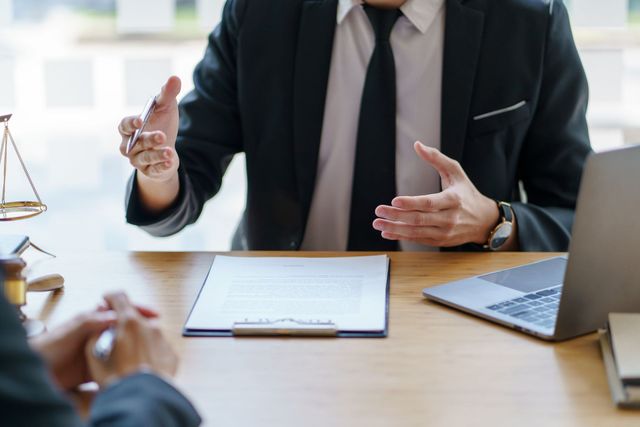 Business meeting at a desk, with a suited person gesturing across papers and a laptop nearby