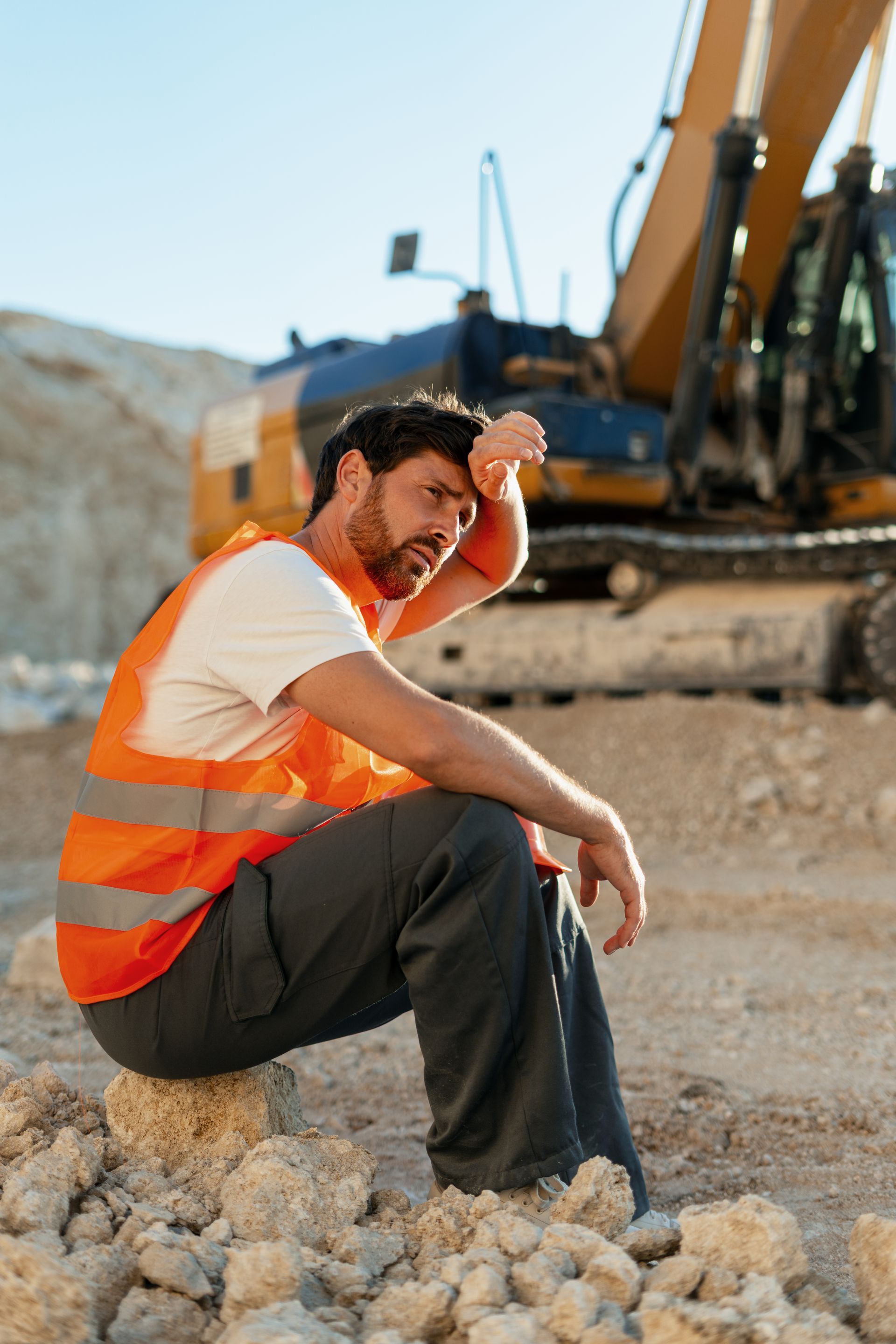 Construction worker in orange safety vest sits on rubble near heavy machinery, shielding his eyes from the sun