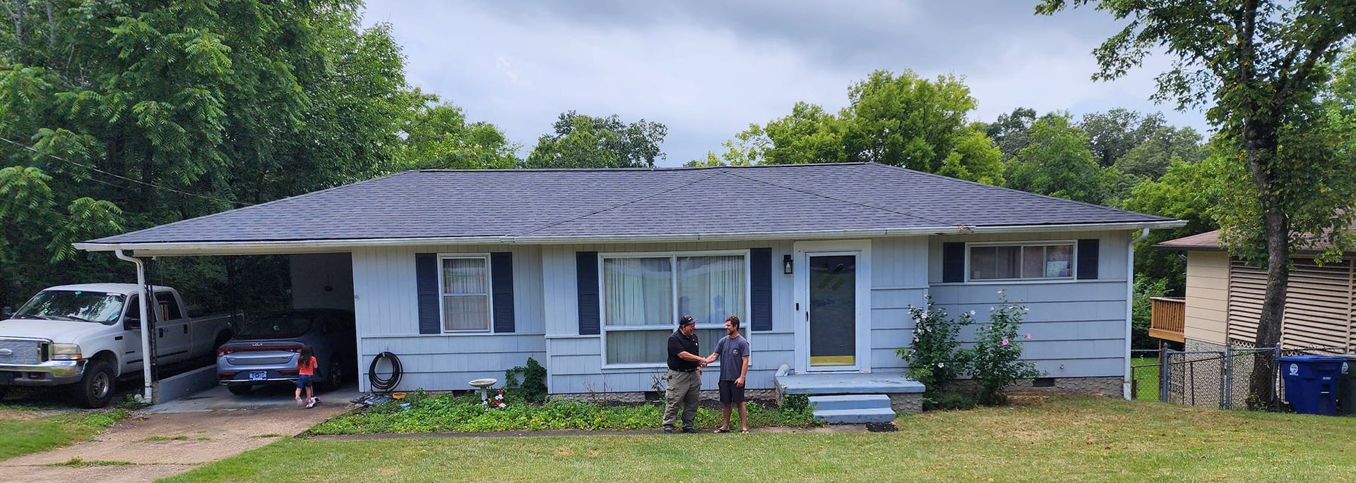 Single-story blue house with a gray roof, front porch, and people standing in the front yard.