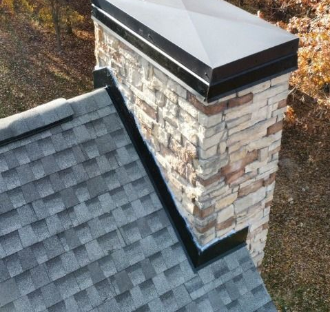 A stone chimney with a black metal cap and flashing, rising from a gray shingled roof against a background of brown foliage.