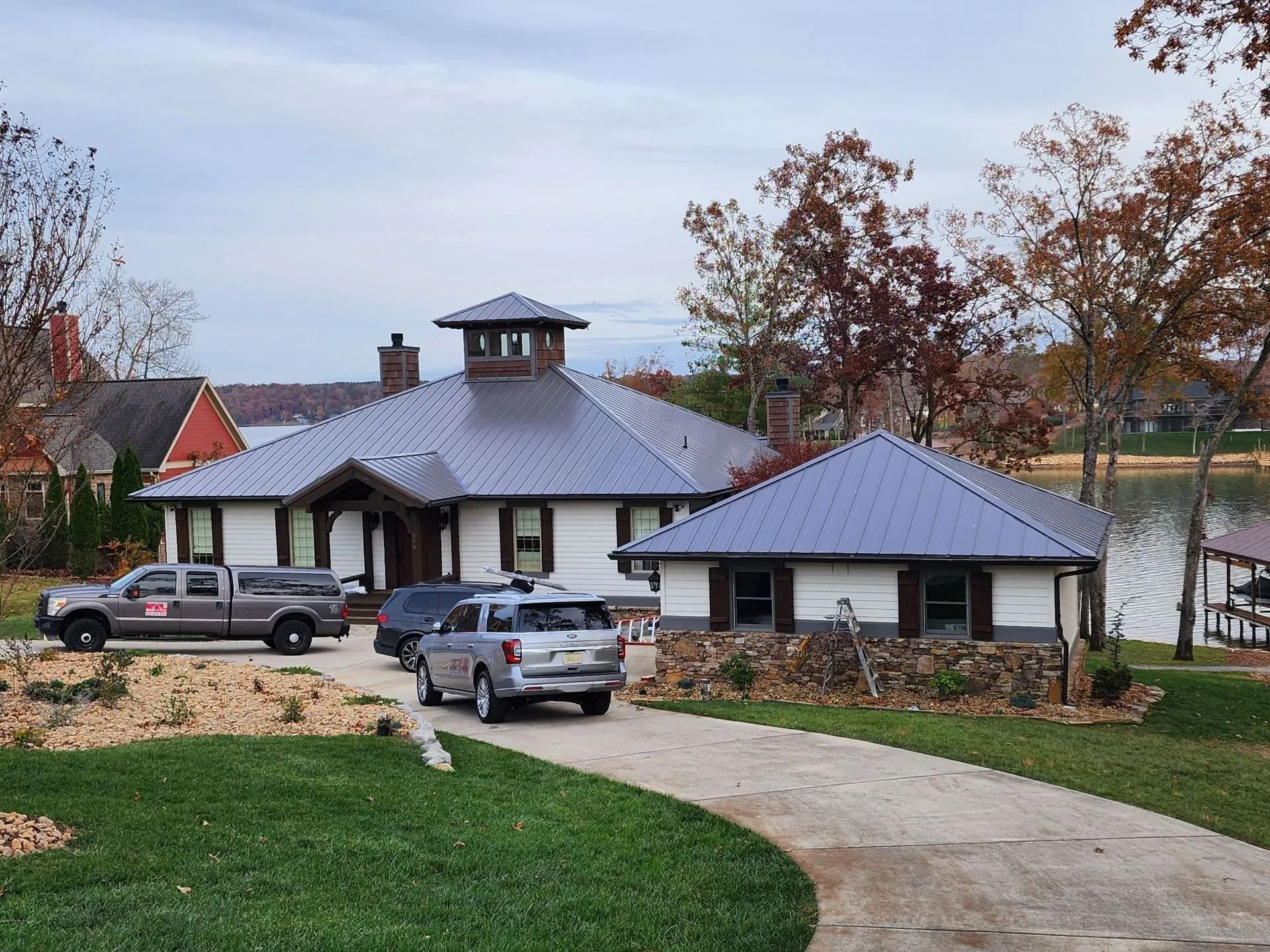 Rural house with gray roof, parked trucks, and curved driveway by a pond under autumn trees