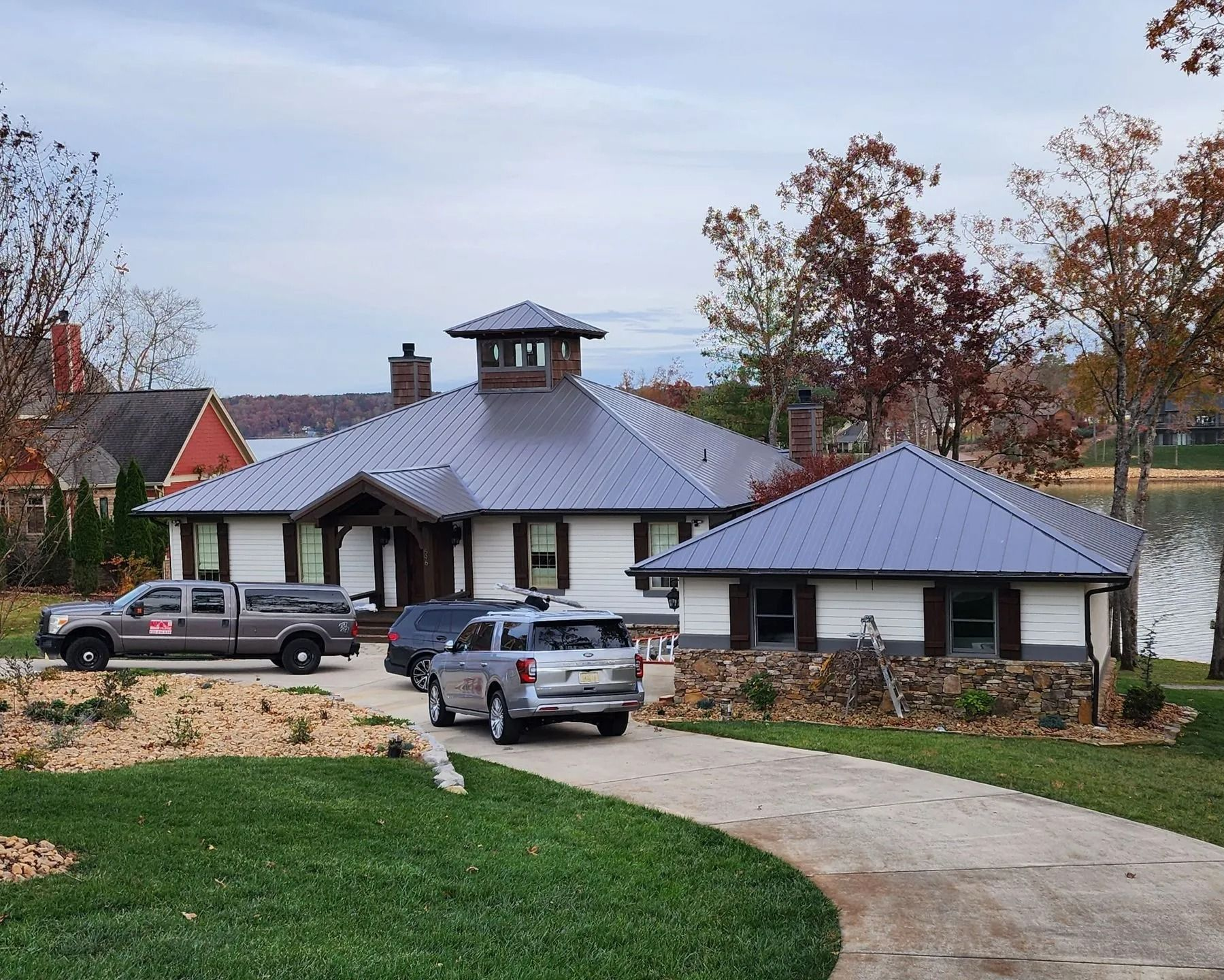 A one-story white home with a dark metal roof and a cupola, featuring a stone base, parked cars, and lakeside trees.