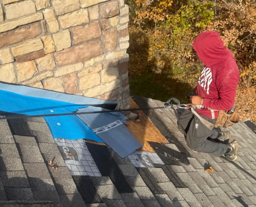 A person in a red hoodie and harness working on a residential roof near a stone chimney.