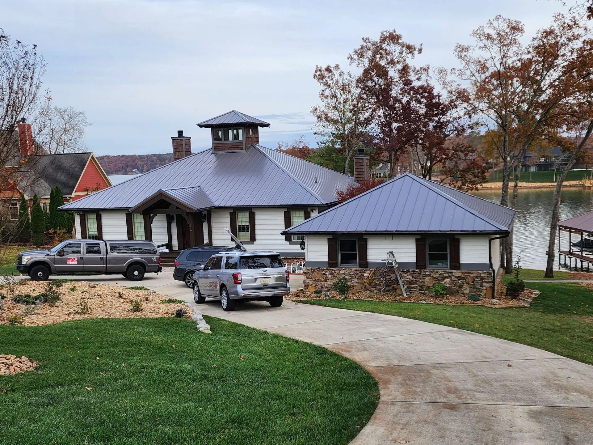 A waterfront house with a metal roof and stone base, featuring a central cupola, parked vehicles, and a paved driveway.