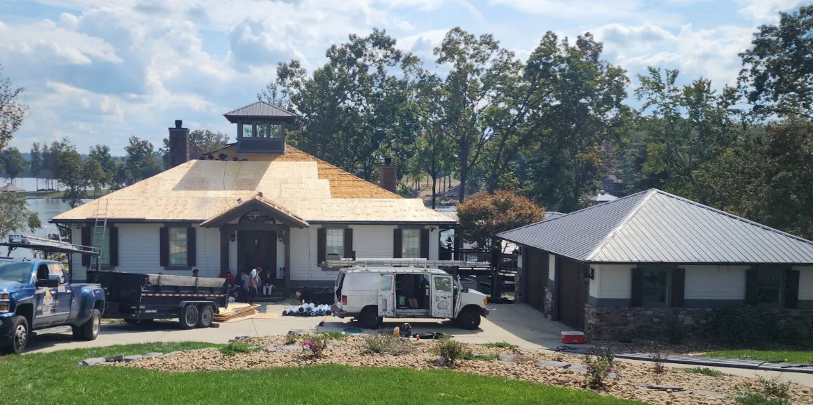 A tan, single-story house with a blue tarp partially covering one side of the dark, worn asphalt shingle roof.