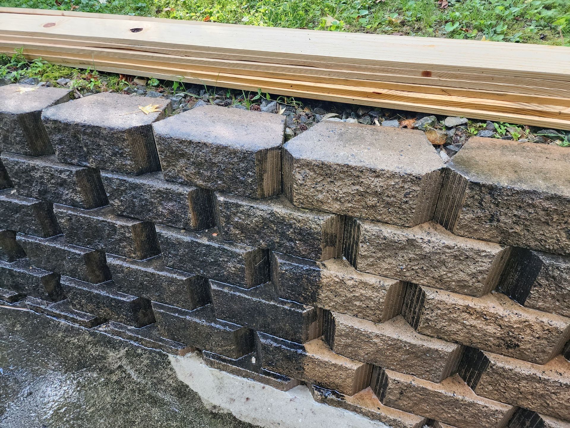A segmented concrete retaining wall, half-covered in dark sealant and half in natural beige stone, sits on a concrete base.