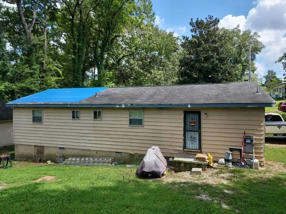A tan, single-story house with a blue tarp partially covering one side of the dark, worn asphalt shingle roof.