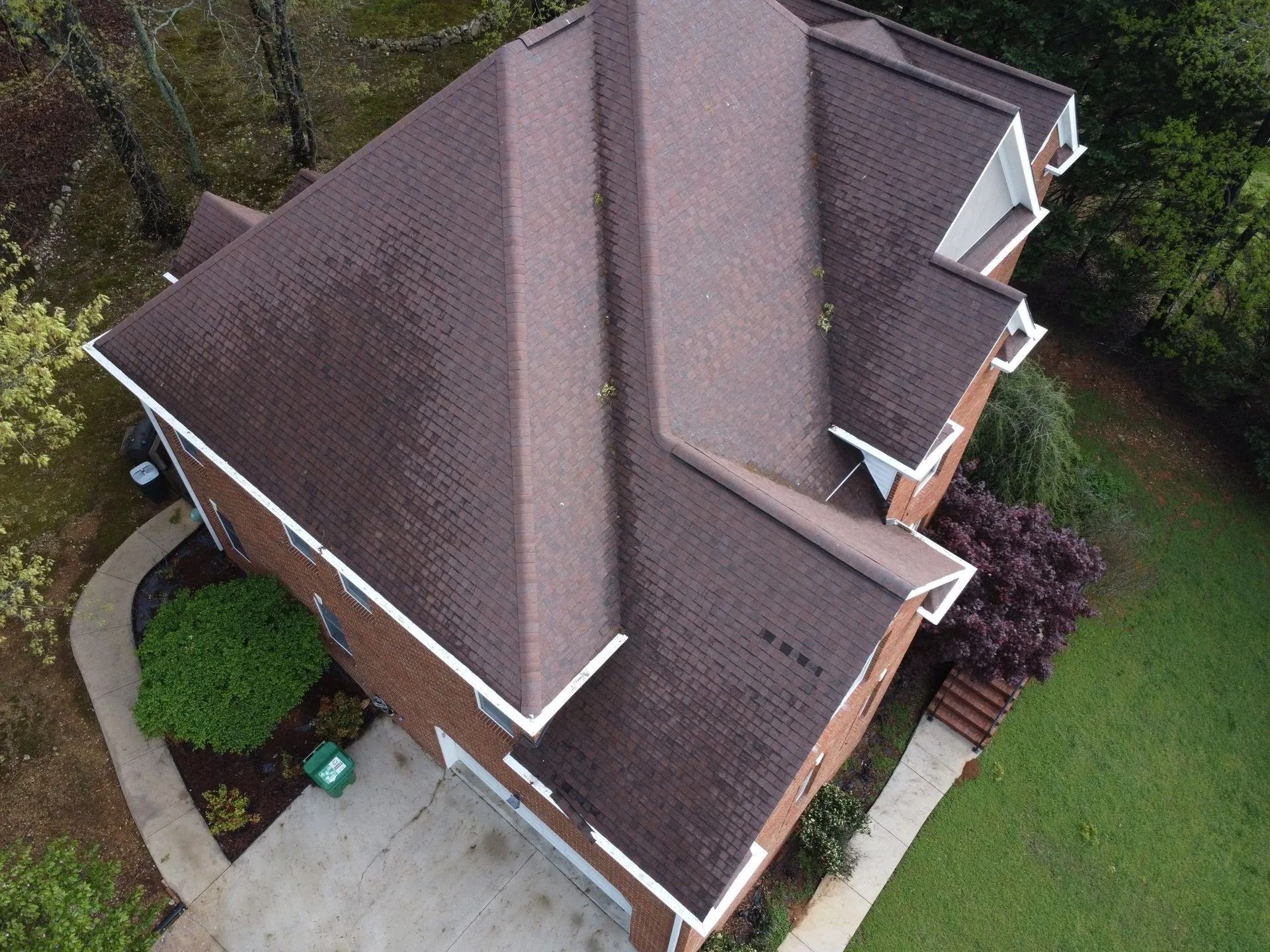 An aerial view of a brick house with a complex dark brown shingled roof, surrounded by a lawn, driveway, and trees.