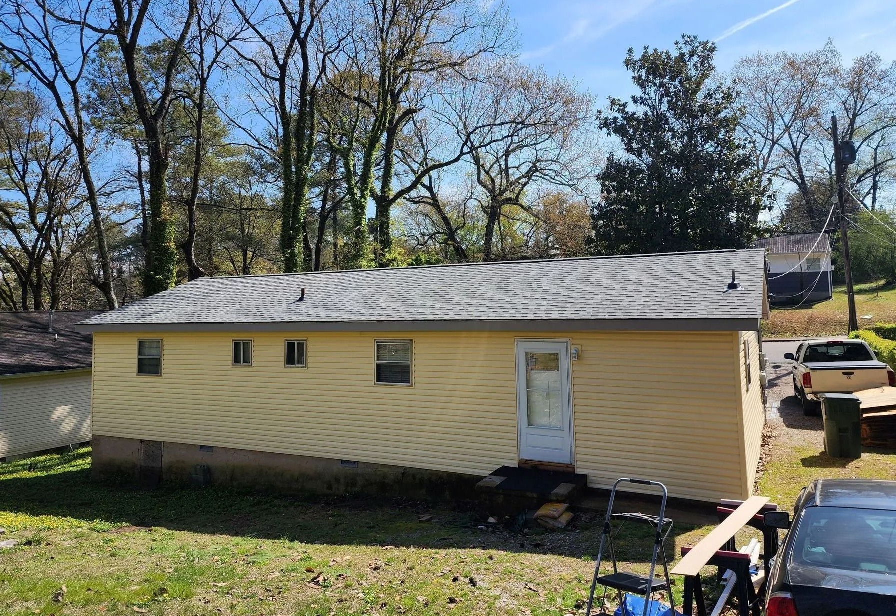 Yellow single-story house with gray roof in a wooded yard on a sunny day