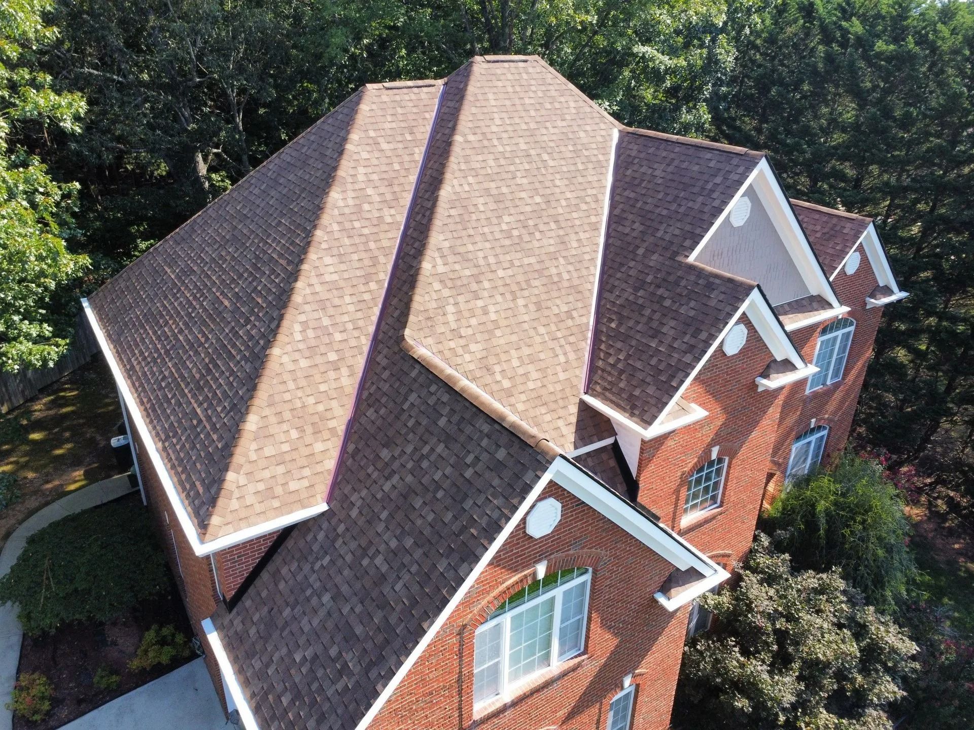 An aerial, high-angle view of a multi-gabled house with a brown shingled roof and red brick siding surrounded by trees.